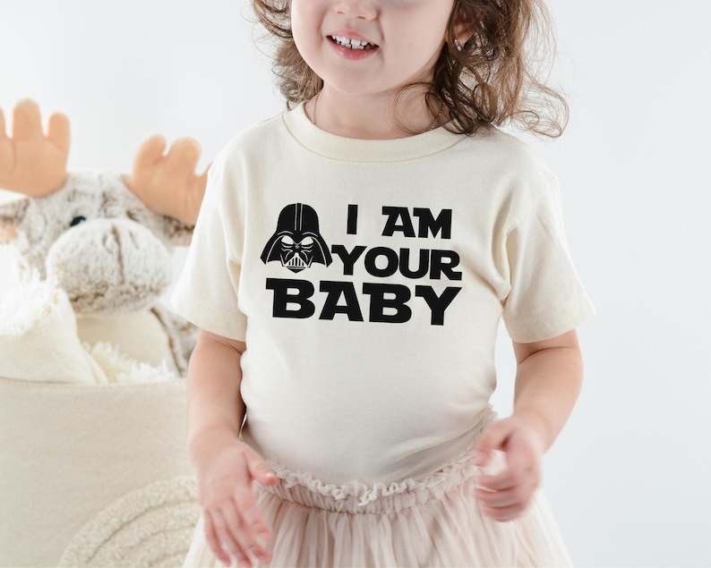 May include: A cream-colored t-shirt with the text "I AM YOUR BABY" in bold black letters, along with a black Darth Vader helmet graphic. The shirt is worn by a child.