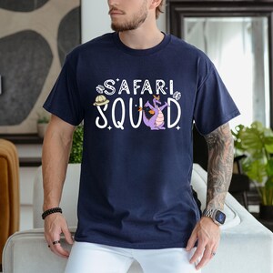 K&ouml;nnte beinhalten: Marineblaues T-Shirt mit dem wei&szlig;en Schriftzug "SAFARI SQUAD", mit einem Cartoon-Drachen und einem Safarihut-Motiv. Der Drache ist lila mit orangefarbenen Akzenten. Das Shirt hat einen Rundhalsausschnitt und kurze &Auml;rmel.