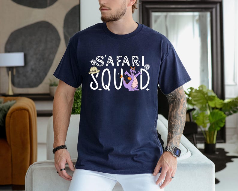 K&ouml;nnte beinhalten: Marineblaues T-Shirt mit dem wei&szlig;en Schriftzug "SAFARI SQUAD", mit einem Cartoon-Drachen und einem Safarihut-Motiv. Der Drache ist lila mit orangefarbenen Akzenten. Das Shirt hat einen Rundhalsausschnitt und kurze &Auml;rmel.