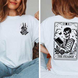 以下が含まれることがあります： 白地に黒と白のグラフィックがプリントされたTシャツ。背面には、本を読んでいる骸骨のイラストと「The Reader」の文字がプリントされている。前面には、バラを持った骸骨の手のイラストがプリントされている。