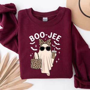 May include: A maroon sweatshirt with a graphic of a ghost wearing sunglasses and a leopard print bow, holding a leopard print bag and a coffee cup. The text "BOO-JEE" is above the ghost. Bats and stars are also in the design.