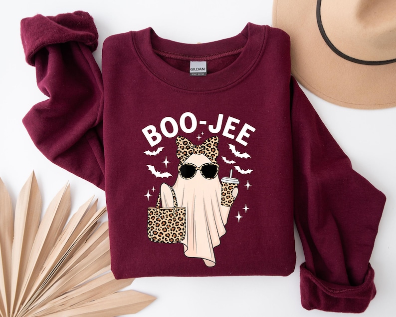 May include: A maroon sweatshirt with a graphic of a ghost wearing sunglasses and a leopard print bow, holding a leopard print bag and a coffee cup. The text "BOO-JEE" is above the ghost. Bats and stars are also in the design.