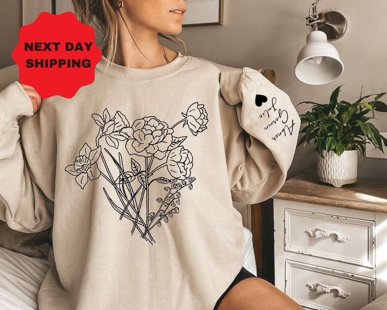 May include: A beige sweatshirt with a black line drawing of flowers on the front. The sleeve has the text "Alice, Grown, True" written in black.