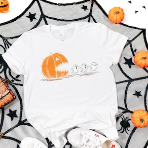 May include: White t-shirt featuring a Halloween-themed graphic. The design shows an orange pumpkin with a carved face chasing three small ghosts. The shirt is surrounded by pumpkins and a spiderweb-like decoration.