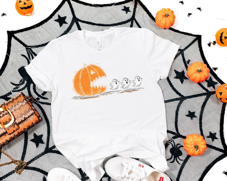 May include: White t-shirt featuring a Halloween-themed graphic. The design shows an orange pumpkin with a carved face chasing three small ghosts. The shirt is surrounded by pumpkins and a spiderweb-like decoration.