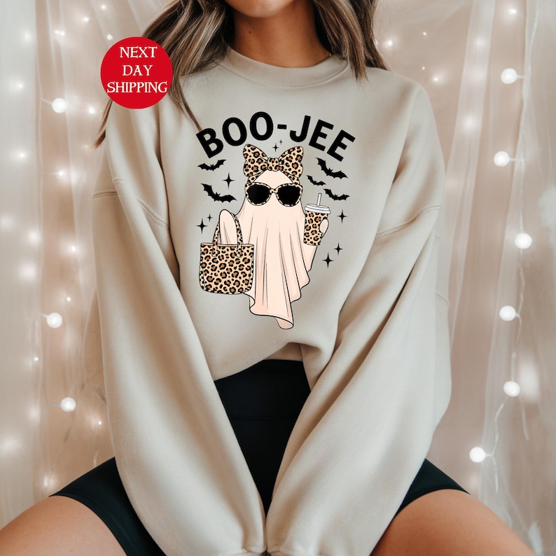 May include: Beige sweatshirt with a Halloween-themed graphic. The design features a ghost wearing sunglasses and a leopard print bow, holding a leopard print tote bag and a coffee cup. The text "BOO-JEE" is above the ghost.