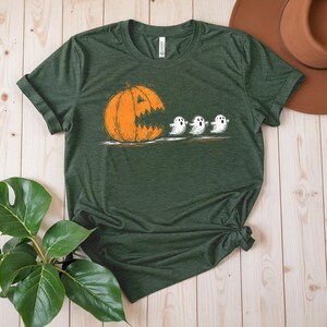 May include: Green t-shirt featuring a Halloween-themed graphic. The design shows an orange pumpkin with a jagged mouth chasing three white ghosts. The shirt is made of a soft material and has a classic crew neck.