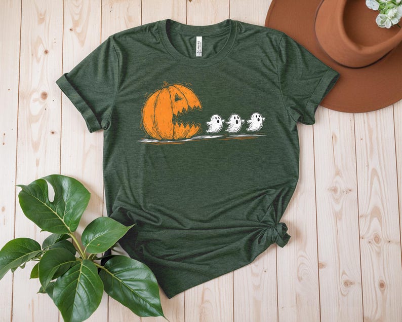 May include: Green t-shirt featuring a Halloween-themed graphic. The design shows an orange pumpkin with a jagged mouth chasing three white ghosts. The shirt is made of a soft material and has a classic crew neck.