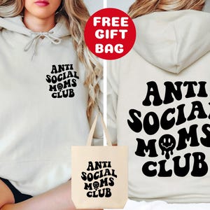 May include: A white hoodie with the text "Anti Social Moms Club" in black, retro-style lettering. The hoodie is shown being worn by a person and comes with a matching tote bag.