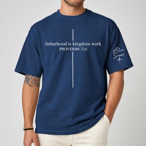 May include: Navy blue t-shirt with the text "fatherhood is kingdom work PROVERBS 22:6" in white. A white cross design is centered on the front. The sleeve has the name "Alex Susan" and a cross. The shirt is a casual style.
