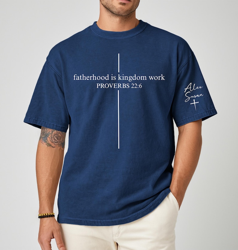 May include: Navy blue t-shirt with the text "fatherhood is kingdom work PROVERBS 22:6" in white. A white cross design is centered on the front. The sleeve has the name "Alex Susan" and a cross. The shirt is a casual style.