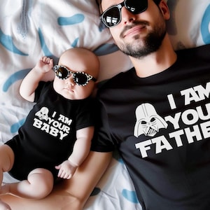 May include: A black t-shirt with the text "I AM YOUR FATHER" and a Darth Vader graphic, next to a baby wearing a black onesie with the text "I AM YOUR BABY" and a Darth Vader graphic. Both are wearing sunglasses.