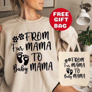 May include: A beige sweatshirt with black text that reads "From Fur Mama to Baby Mama" and two paw prints. A beige tote bag with the same text and paw prints is also shown.