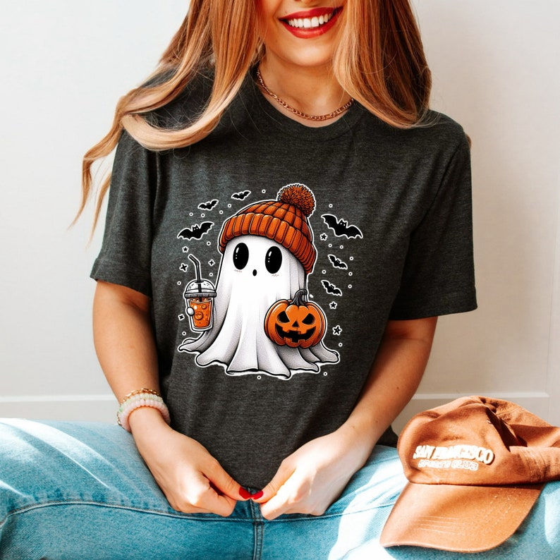 Może przedstawiać: Szary t-shirt z nadrukiem kresk&oacute;wkowego ducha w pomarańczowo-brązowej czapce, trzymającego dynię i kubek pomarańczowego napoju. Duch otoczony jest nietoperzami i gwiazdami.
