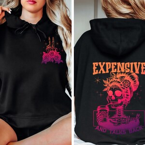 May include: Black hoodie with a pink and orange gradient design. The front features a skeleton hand making a peace sign with roses. The back features a skeleton with a bun, drinking from a cup with the text "Expensive and Talks Back".