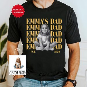 Puede incluir: Camiseta negra con el texto dorado "EMMA'S DAD" y una foto en blanco y negro de una ni&ntilde;a. La camiseta tambi&eacute;n incluye el texto "EST. 2020". Una peque&ntilde;a foto con el texto "CUSTOM PHOTO" est&aacute; en la esquina inferior izquierda.