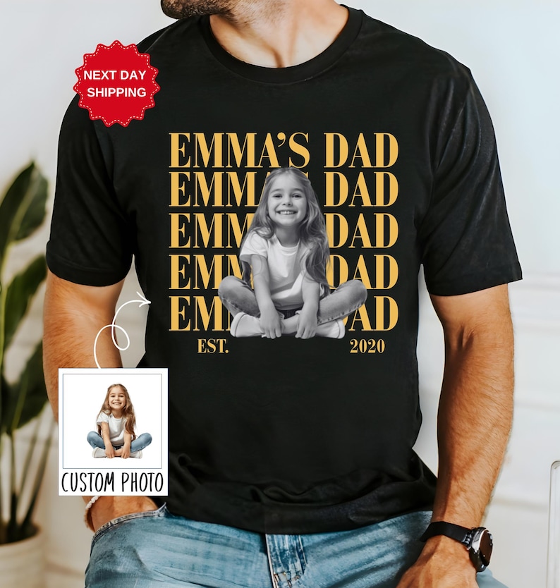 Puede incluir: Camiseta negra con el texto dorado "EMMA'S DAD" y una foto en blanco y negro de una ni&ntilde;a. La camiseta tambi&eacute;n incluye el texto "EST. 2020". Una peque&ntilde;a foto con el texto "CUSTOM PHOTO" est&aacute; en la esquina inferior izquierda.