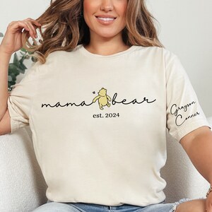 May include: A white t-shirt with the text "mama bear est. 2024" and a cartoon Winnie the Pooh bear. The text "Grayson Conner" is on the sleeve of the shirt.