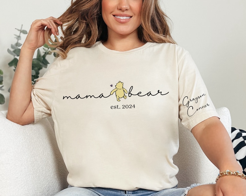 May include: A white t-shirt with the text "mama bear est. 2024" and a cartoon Winnie the Pooh bear. The text "Grayson Conner" is on the sleeve of the shirt.