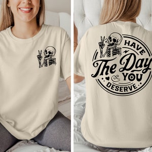 May include: A beige t-shirt with a skeleton graphic on the front and the text "Have The Day You Deserve" with a skeleton graphic on the back.