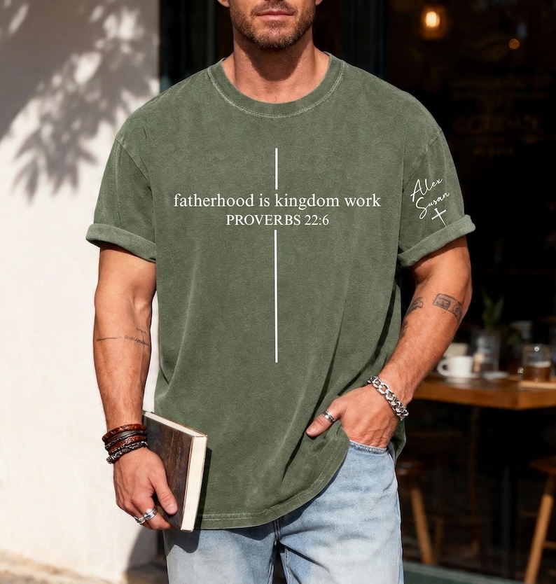 May include: Olive green t-shirt with the text "fatherhood is kingdom work PROVERBS 22:6" in white. A white cross graphic is centered below the text. The shirt has rolled-up sleeves and a small signature with a cross on the right sleeve.