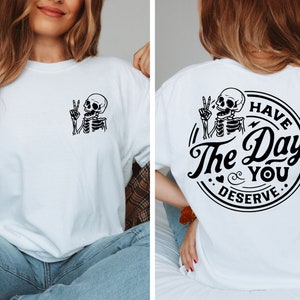 May include: White t-shirt with a black skeleton graphic on the front and a black circle graphic on the back that says "Have the Day You Deserve".