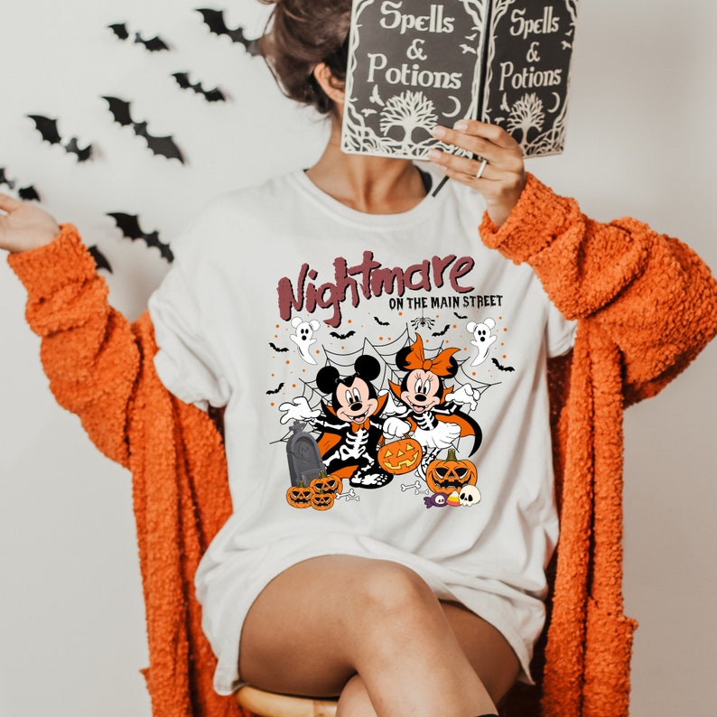 May include: White t-shirt featuring a graphic of Mickey and Minnie Mouse dressed in Halloween costumes. The text "Nightmare on the Main Street" is printed on the shirt.