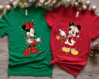 以下が含まれることがあります： クリスマスをテーマにした2枚のTシャツ。1つは、プレゼントを持ってクリスマスライトに包まれたミニーマウスが描かれた緑色です。もう1つは、サンタの帽子をかぶり、オーナメントを持ったミッキーマウスが描かれた赤色で、こちらもライトで包まれています。お祝いの服。