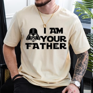 May include: Beige short-sleeve t-shirt featuring a black graphic of Darth Vader's helmet and the text "I AM YOUR FATHER". The design is centered on the front of the shirt. The shirt is made of a soft, comfortable material.
