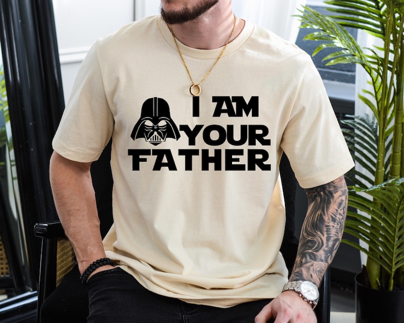 May include: Beige short-sleeve t-shirt featuring a black graphic of Darth Vader's helmet and the text "I AM YOUR FATHER". The design is centered on the front of the shirt. The shirt is made of a soft, comfortable material.