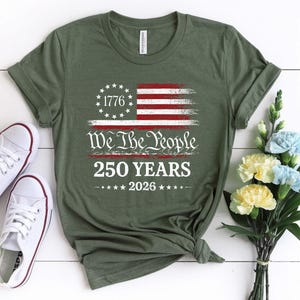 May include: An olive green short-sleeve t-shirt featuring a distressed American flag graphic with a circle of stars, the text 1776, We The People, 250 YEARS, and 2026. The shirt has a crew neckline and is displayed with white sneakers and flowers.
