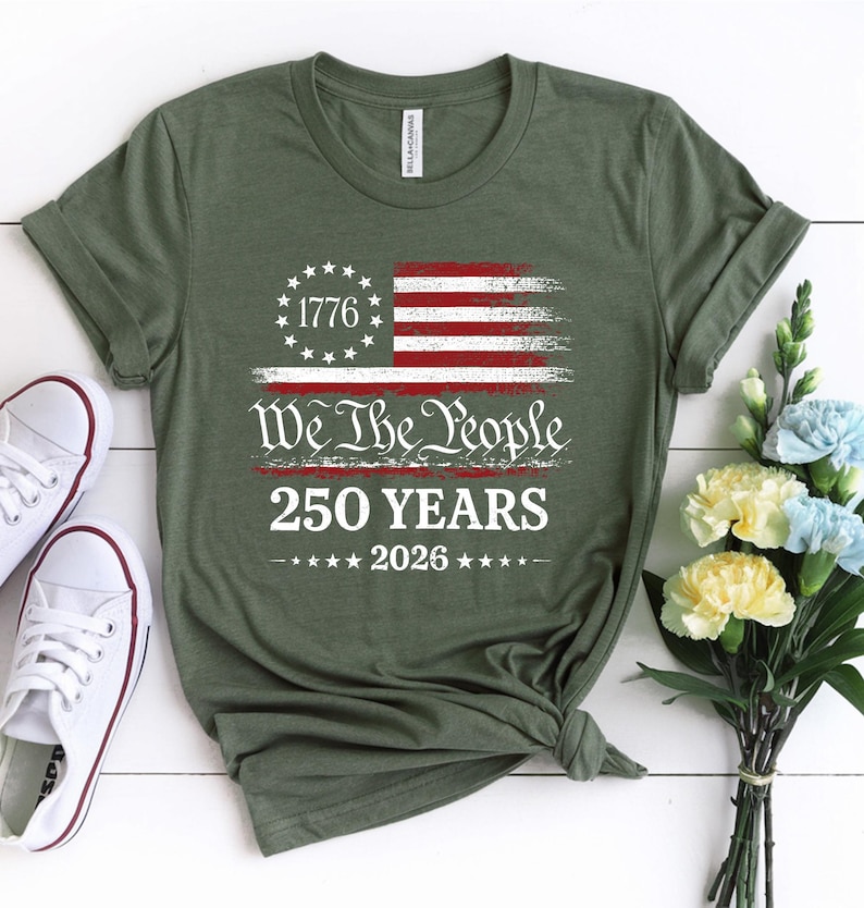 May include: An olive green short-sleeve t-shirt featuring a distressed American flag graphic with a circle of stars, the text 1776, We The People, 250 YEARS, and 2026. The shirt has a crew neckline and is displayed with white sneakers and flowers.