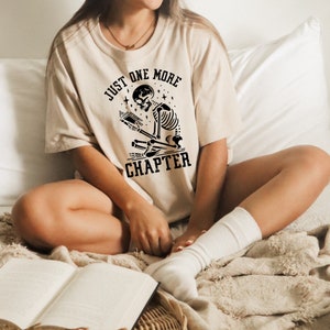 May include: A beige t-shirt with a black graphic of a skeleton reading a book. The graphic includes the text "Just One More Chapter".