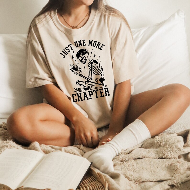 May include: A beige t-shirt with a black graphic of a skeleton reading a book. The graphic includes the text "Just One More Chapter".