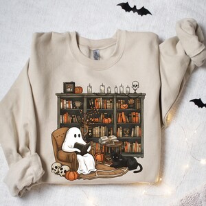 May include: A beige sweatshirt with a graphic of a ghost reading a book in a chair next to a bookshelf filled with books and pumpkins. The bookshelf is decorated with candles and a skull.