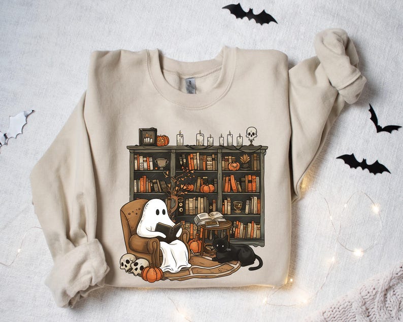 May include: A beige sweatshirt with a graphic of a ghost reading a book in a chair next to a bookshelf filled with books and pumpkins. The bookshelf is decorated with candles and a skull.