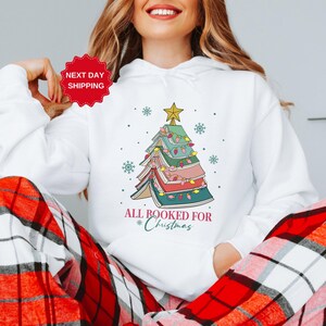 Puede incluir: Sudadera blanca con un &aacute;rbol de Navidad hecho de libros y el texto "All Booked for Christmas".