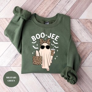 May include: Military green sweatshirt with the text "BOO-JEE" and a graphic of a ghost wearing sunglasses and a leopard print bow, holding a purse and a coffee cup. The design includes bats and stars.