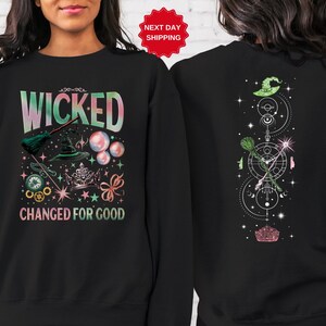 Puede incluir: Sudadera negra con las palabras "WICKED CHANGED FOR GOOD" en una fuente colorida y caprichosa. El dise&ntilde;o frontal incluye un sombrero de bruja, una escoba y burbujas. La parte trasera presenta un sombrero de bruja, una escoba y un dise&ntilde;o geom&eacute;trico.