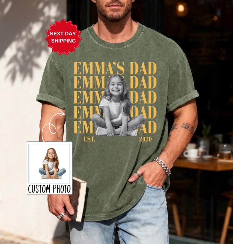 Puede incluir: Camiseta verde oliva con el texto "EMMA'S DAD" repetido verticalmente y una foto en blanco y negro de una ni&ntilde;a. La camiseta tambi&eacute;n tiene el texto "EST. 2020". Una peque&ntilde;a imagen con el texto "CUSTOM PHOTO" est&aacute; en la esquina inferior izquierda.