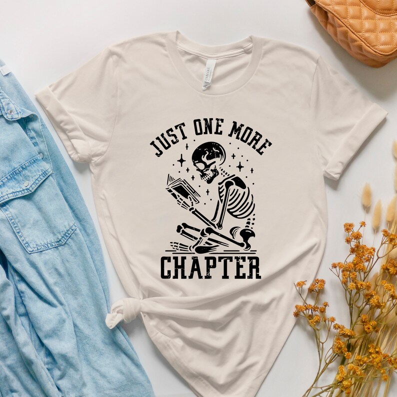May include: A beige t-shirt with a black graphic of a skeleton reading a book. The text "Just One More Chapter" is printed below the skeleton.