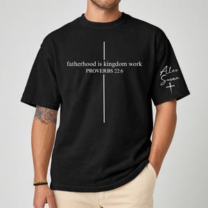 May include: Black t-shirt with the text "fatherhood is kingdom work PROVERBS 22:6" and a white cross design. The sleeve features the signature "Alex Susan" and a cross. The shirt is a casual wear item.