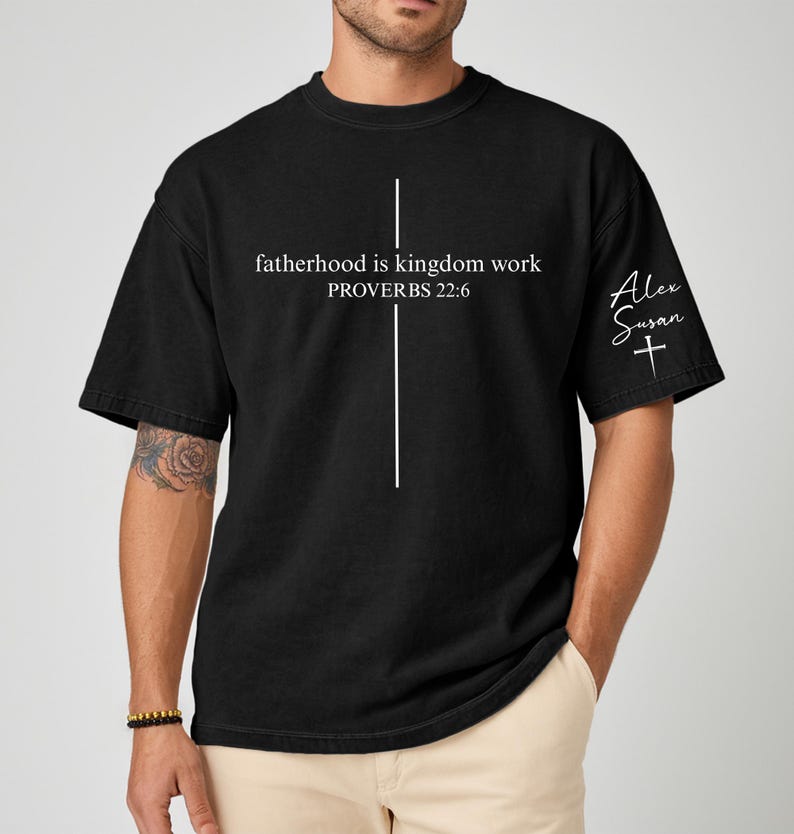 May include: Black t-shirt with the text "fatherhood is kingdom work PROVERBS 22:6" and a white cross design. The sleeve features the signature "Alex Susan" and a cross. The shirt is a casual wear item.