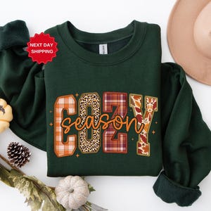 May include: A dark green sweatshirt with the words "COZY season" in a plaid, leopard print, and floral design. The sweatshirt has a crew neck and long sleeves. A red sticker that says "NEXT DAY SHIPPING" is visible.