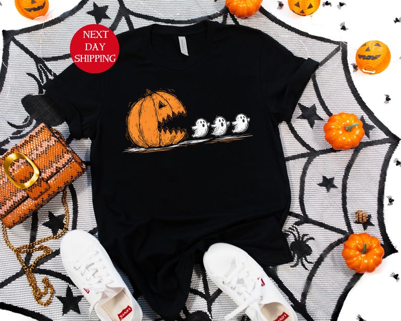 May include: Black t-shirt featuring a Halloween-themed graphic. The design shows an orange pumpkin with a carved face chasing three white ghosts. The shirt is displayed with white sneakers and a decorative purse.