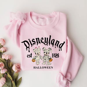 May include: Pink sweatshirt with a black graphic of Mickey Mouse and Minnie Mouse dressed as mummies. The graphic says "Disneyland est 1928 HALLOWEEN".