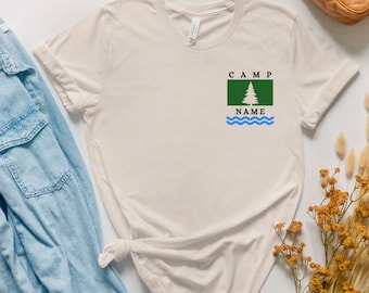 May include: A beige t-shirt with a green and blue camp flag design. The flag has a pine tree and the text "CAMP NAME".
