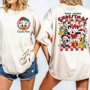 Op de afbeelding: Een cr&egrave;mekleurig T-shirt met een kerstthema met Mickey Mouse, Minnie Mouse, Donald Duck, Goofy en Pluto. De tekst "Very Merry Christmas" staat op het shirt gedrukt.