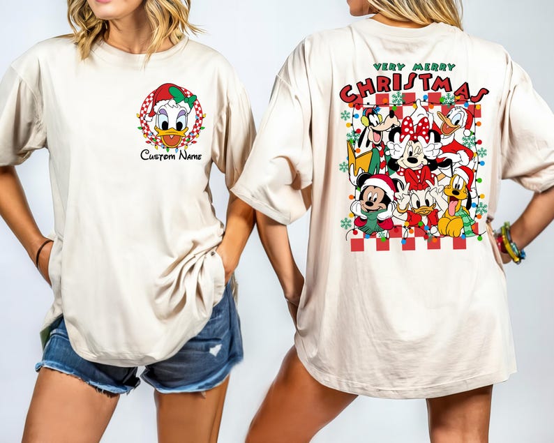 Op de afbeelding: Een cr&egrave;mekleurig T-shirt met een kerstthema met Mickey Mouse, Minnie Mouse, Donald Duck, Goofy en Pluto. De tekst "Very Merry Christmas" staat op het shirt gedrukt.