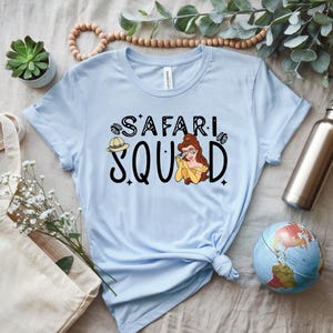K&ouml;nnte beinhalten: Hellblaues T-Shirt mit dem Schriftzug "SAFARI SQUAD" in schwarzer Schrift und einer Cartoon-Illustration einer Frau mit Brille und gelbem Kleid. Ein Safarihut ist ebenfalls abgebildet.